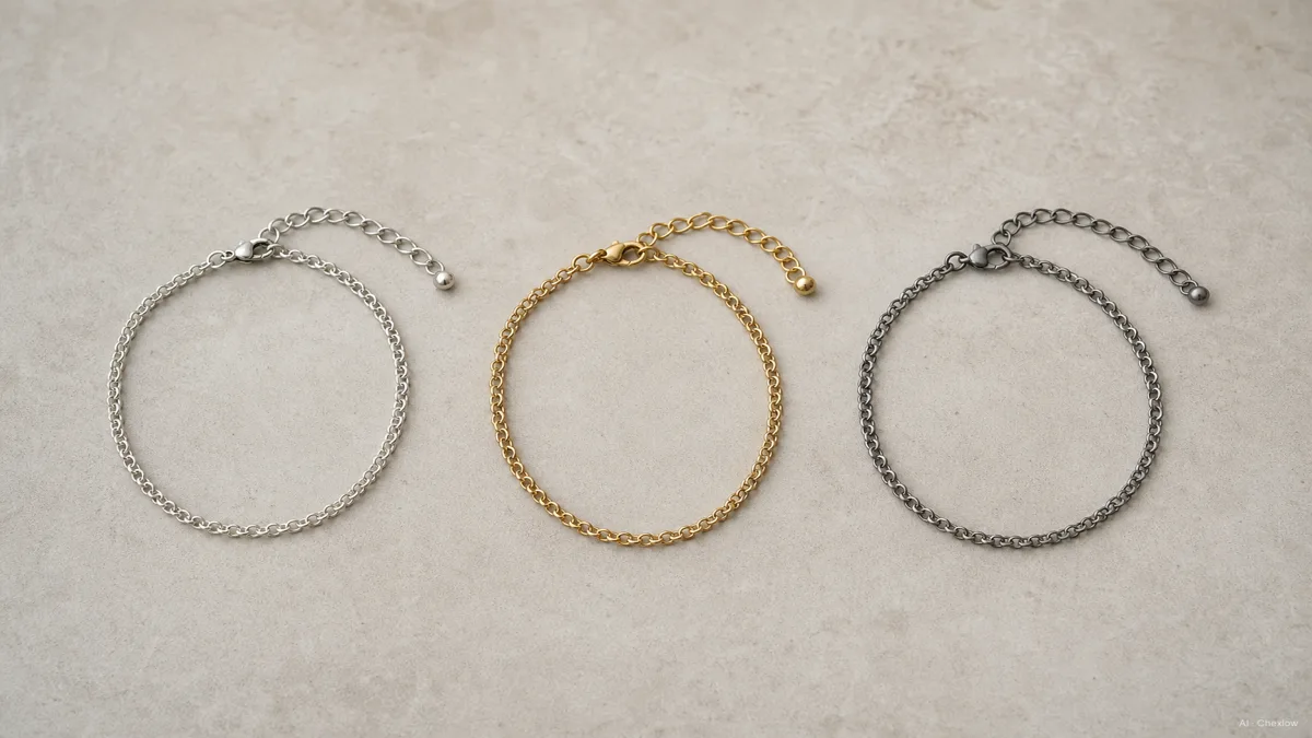 Image: three anklets laid flat side by side on a neutral surface, a silver chain, a gold-toned chain, and a matte steel chain, soft studio lighting