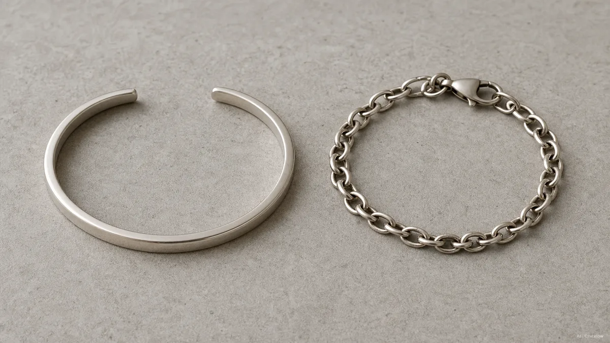 Image: a rigid open-ended cuff bracelet and a flexible chain bracelet with clasp laid side by side on a neutral surface, showing the structural gap versus the linked chain, natural light
