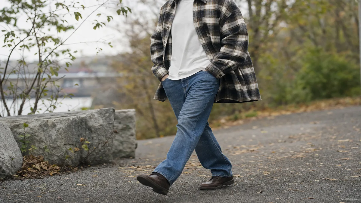 Image: a person wearing an open heavyweight plaid flannel shacket over a plain t-shirt and jeans, walking outdoors on a cool overcast day, relaxed roomy fit visible