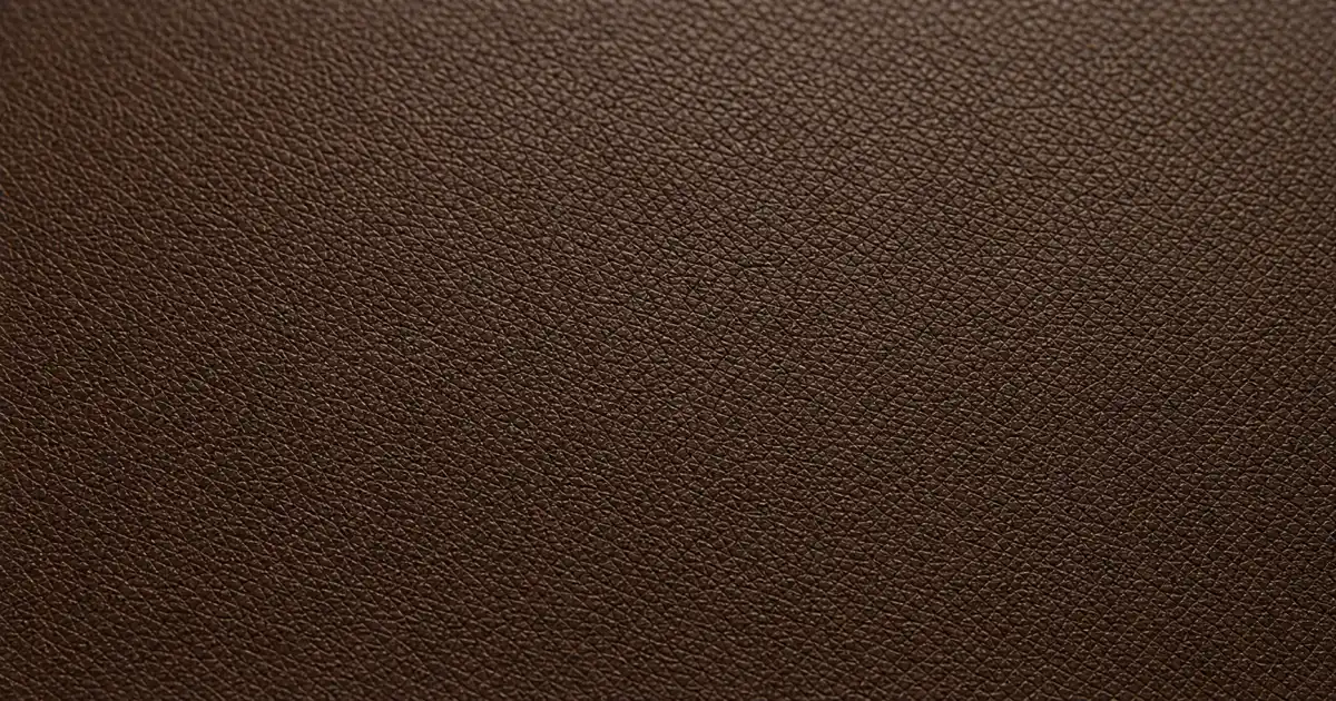 A piece of corrected grain leather laid flat on a matte oak desk in soft natural daylight, showing a perfectly uniform embossed surface pattern, no visible natural pores, slightly glossy due to artificial coating (AI generated illustration)