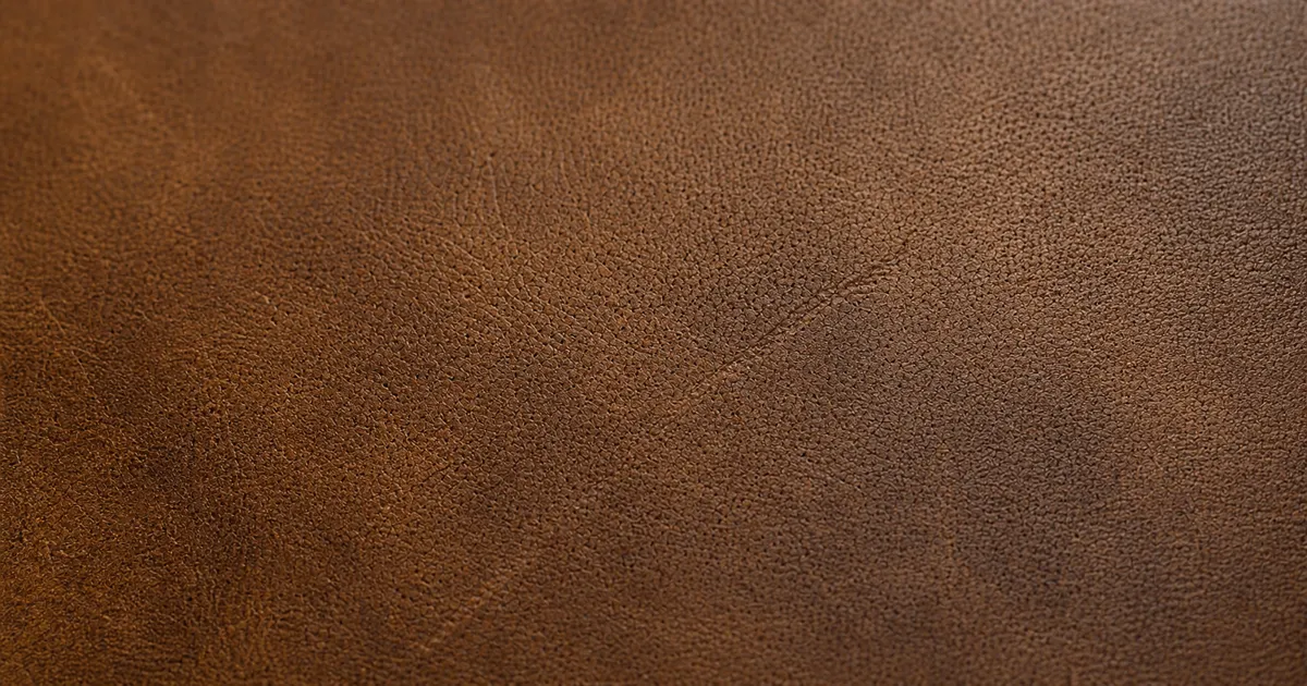 A piece of full grain leather laid flat on a matte oak desk in soft natural daylight, showing visible natural pores, fine tonal variation, and a small natural scar — the outermost layer of the hide intact (AI generated illustration)
