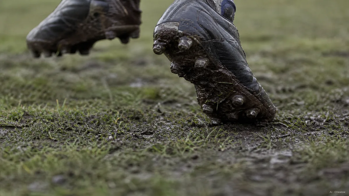 Image: A close-up of a player's cleats mid-stride on a muddy soccer pitch, mud visible around the studs, natural overcast light, no visible brand marks or logos